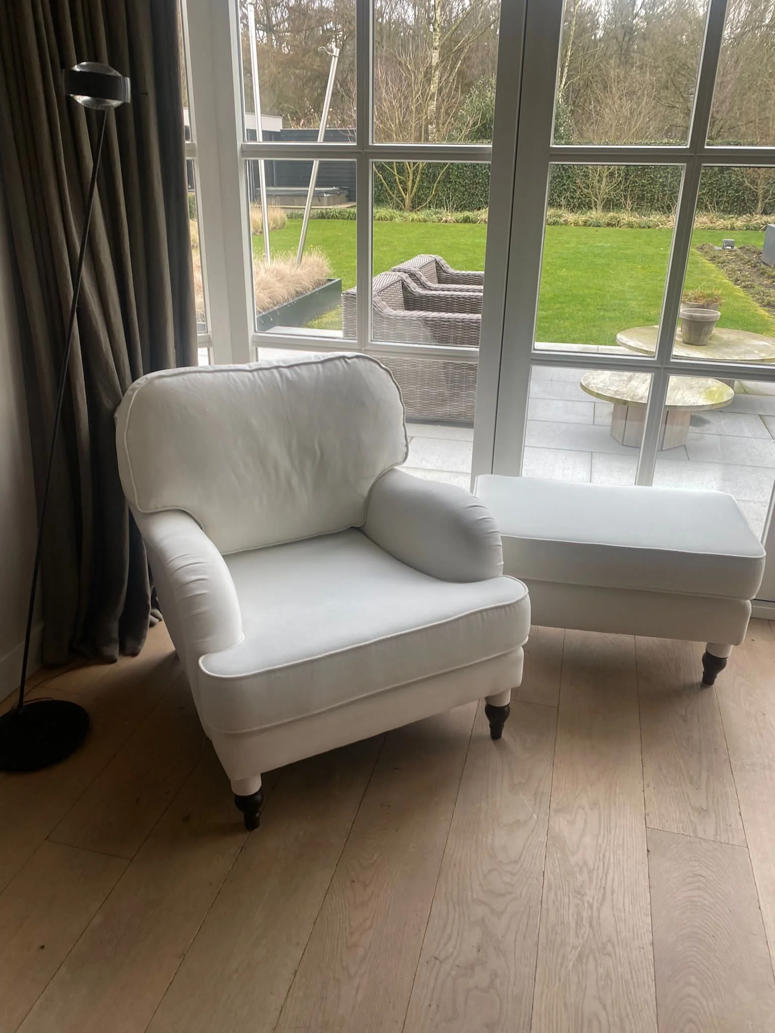 Witte gestoffeerde fauteuil met poef bij een groot raam met uitzicht op de tuin
