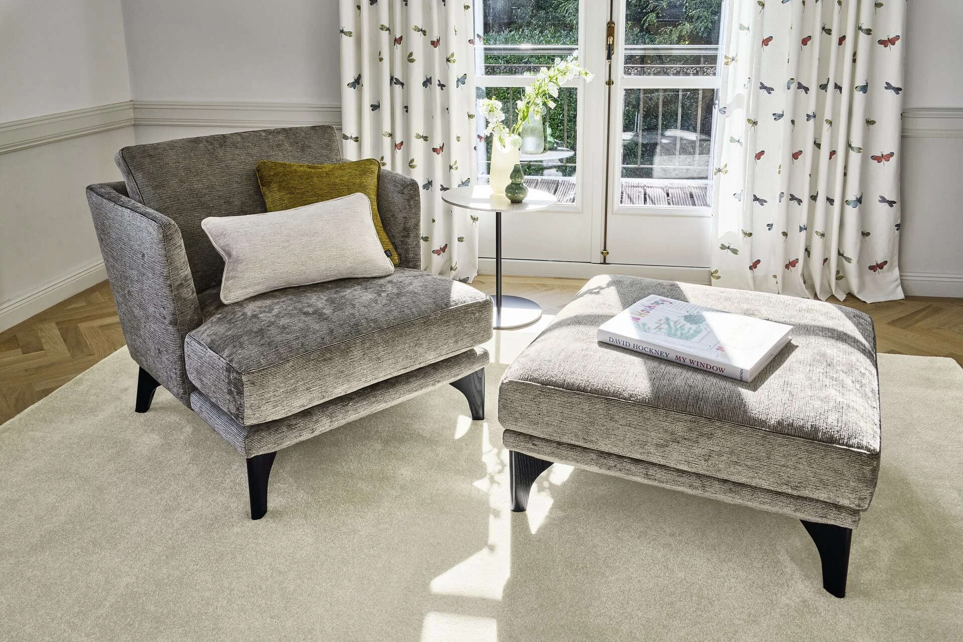 Grijze velvet fauteuil met poef in een lichte zonnige woonkamer met visgraat parket